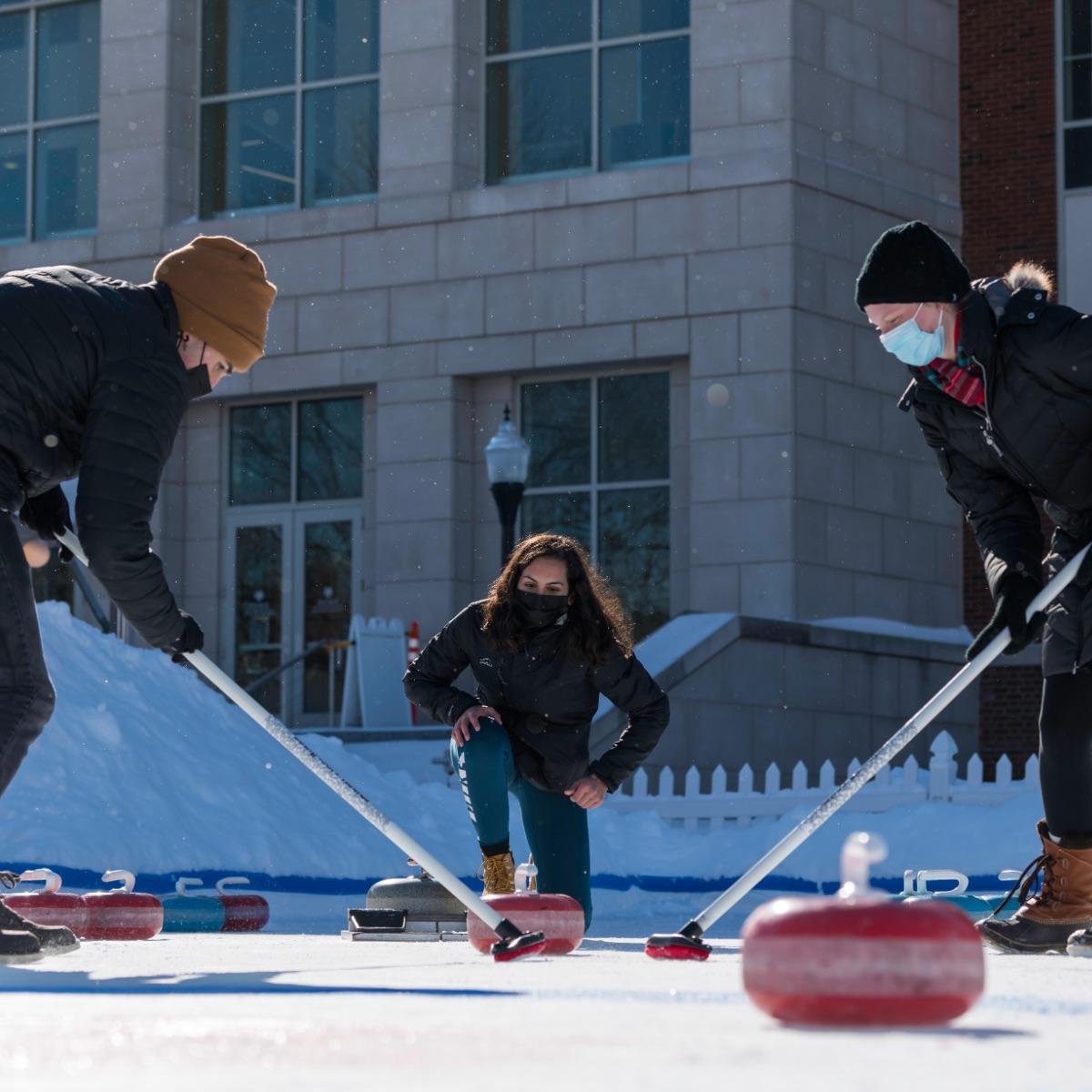 Three people wearing masks participate in curling in winter. One is on their knee, and two are sweeping